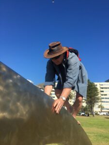 A photo of Jeremy climbing cautiously on top of an arm of the Sunglasses sculpture, on the Sea Point promenade.